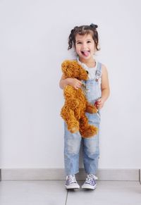 Smiling girl with teddy bear standing against wall at home