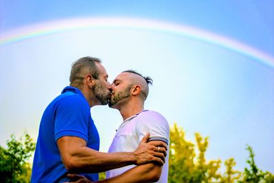 Side view of mid adult gay couple kissing against rainbow in sky