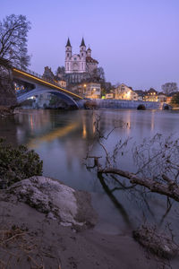 Illuminated bridge over river by buildings against sky at dusk