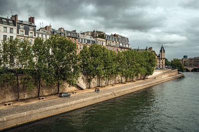 Trees by river against buildings in city against sky