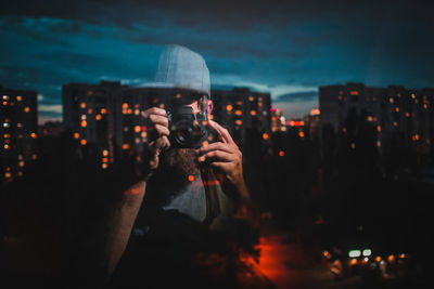 Reflection man photographing illuminated buildings in city on window at night