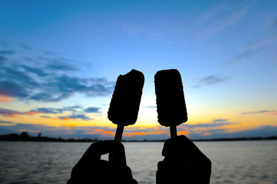 Close-up of silhouette hands having popsicles on beach against sky during sunset