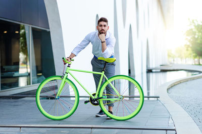 Young bearded man leaning on bicycle seat while looking at camera