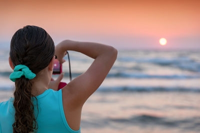 Rear view of girl photographing while standing at beach during sunset