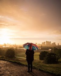 Rear view of woman with umbrella standing on field against cloudy sky during sunset