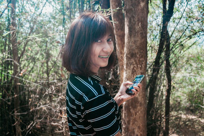 Portrait of young woman using phone while standing on tree trunk