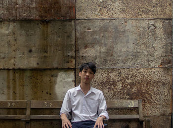 Portrait of young man sitting on bench against wall