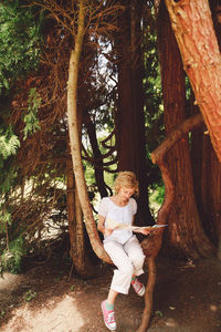 Full length of young woman reading map while sitting on branch of tree