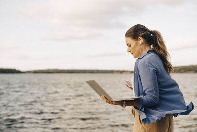 Confident mature woman using laptop while talking on mobile phone by lake