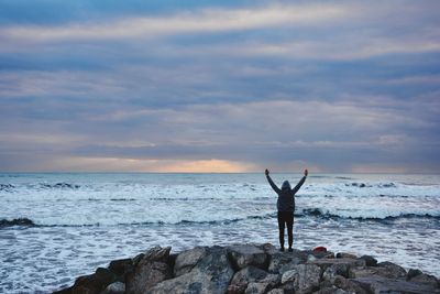 Rear view of person with arms raised standing on rock by mediterranean sea against cloudy sky