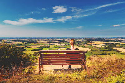 Rear view of woman sitting on bench by agricultural field against sky