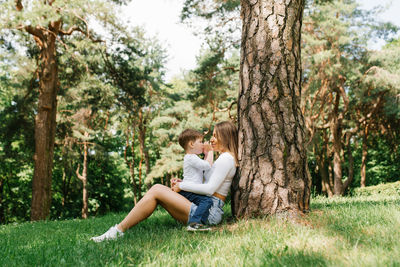 Young caucasian mom and her three-year-old son have fun sitting in the park near a tree on the grass