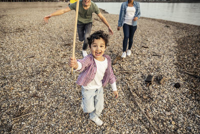 Playful boy holding sticks while playing with parents in background over pebble