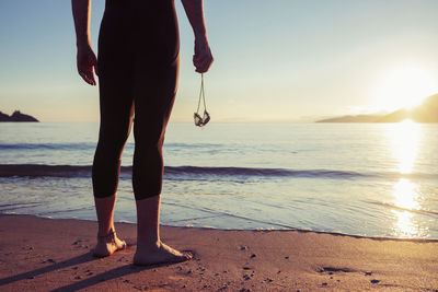 Back view of crop faceless swimmer in swimsuit and with goggles standing on wet beach at sunset over sea