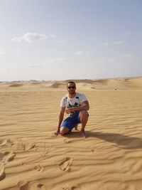 Portrait of man kneeling on sand in desert against sky