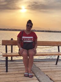 Portrait of woman sitting on bench against sea during sunset