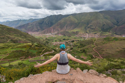 Rear view of woman with arms outstretched sitting on mountain against cloudy sky