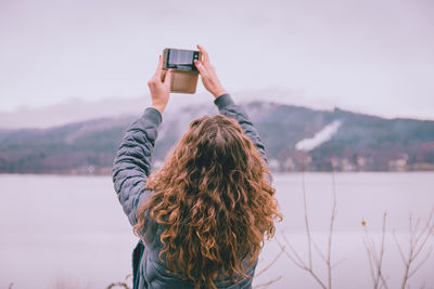 Rear view of man photographing while standing by lake against sky
