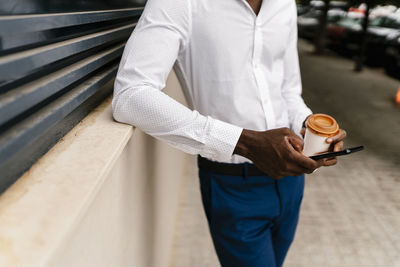 Young male professional holding reusable cup and using smart phone while leaning on surrounding wall