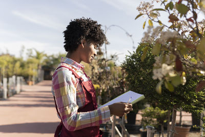 Young gardener holding book standing near plants in nursery