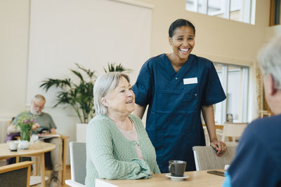 Cheerful young female nurse standing by senior woman while looking at man in retirement home