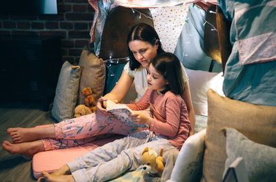 Mother and daughter having a pajama party reading a book in a diy tent