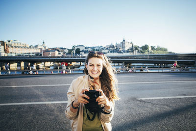 Portrait of happy tourist holding camera on bridge against clear sky