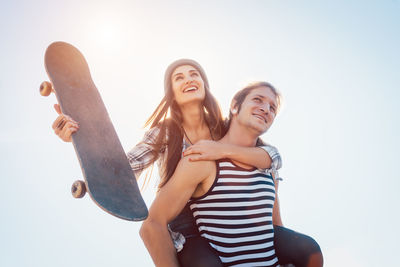 Low angle view of man piggybacking cheerful girlfriend with skateboard against clear sky