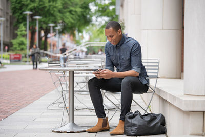 Full length of young man using phone while sitting on footpath