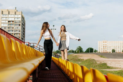 Two teenage girls walk together through the stands of the school stadium, talking, holding hands