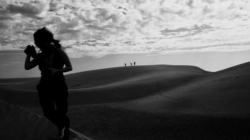 Woman holding camera walking on desert against cloudy sky
