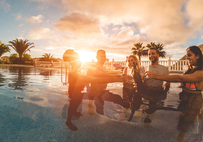 Friends toasting beer bottles and glasses in swimming pool against sky during sunset