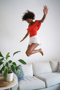 Happy black woman with curly hair having fun jumping dancing on the couch at home