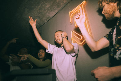 Cheerful young dj gesturing amidst friends dancing at illuminated nightclub