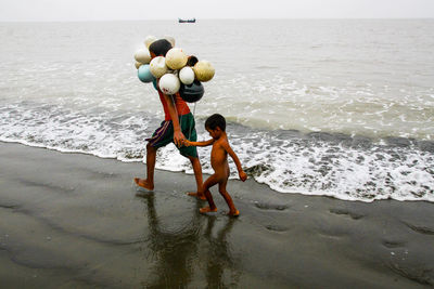 Boy carrying buoys while holding naked sibling hand at beach