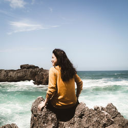 Rear view of young woman looking away while sitting on rock by sea against sky