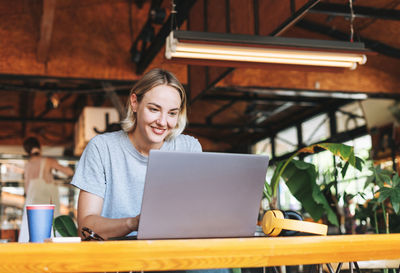 Young smiling blonde woman freelancer in yellow headphones working on laptop on the table at cafe