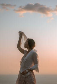 Woman holding fabric while standing against sky during sunset
