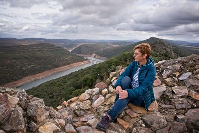 Man sitting on rock looking at mountains against sky