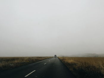 Road passing through field against sky during foggy weather