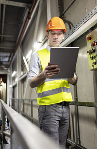 Worker in reflective clothing using tablet pc by railing in warehouse