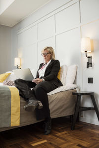 Businesswoman sitting on bed using laptop against wall in hotel room