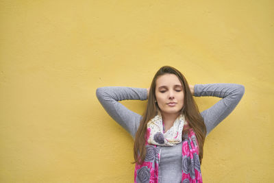 Young woman with eyes closed hand hands behind head standing against yellow wall