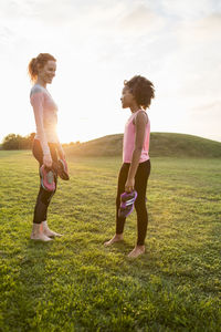 Side view of mother and daughter holding sports shoes standing on grass at park against sky during sunset