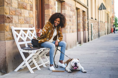 Happy woman sitting with dog taking selfie on mobile phone at sidewalk in city