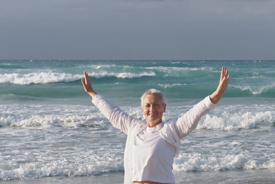 Happy caucasian senior woman looking at camera, with outstretched arms, against backdrop of ocean