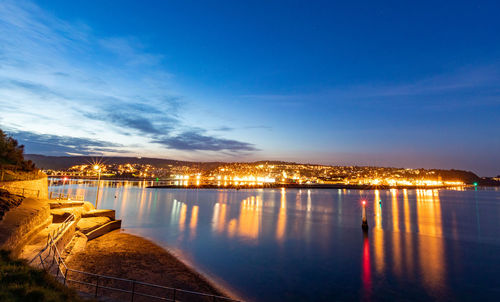 Illuminated city by river against sky at dusk