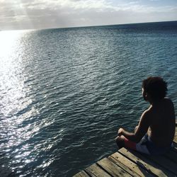 Shirtless man sitting on pier over sea against sky