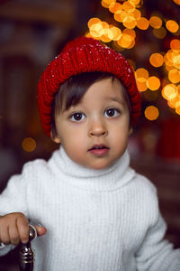 Child in a white sweater and a red knitted hat stands next to the christmas tree at home