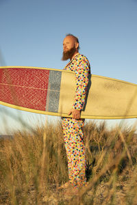 Man in colorful suit with surfboard looking away while standing against sky